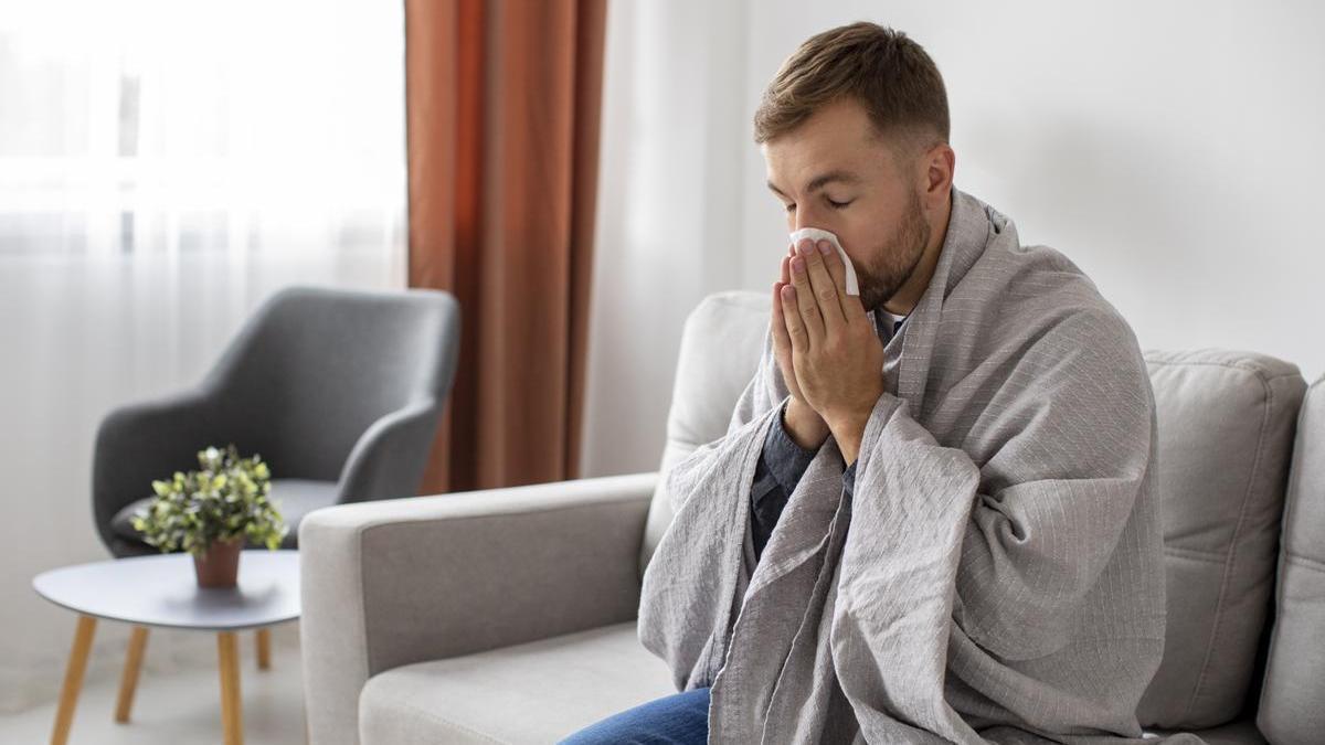 Hombre con gripe se suena la nariz en el sofá mientras se tapa con una manta