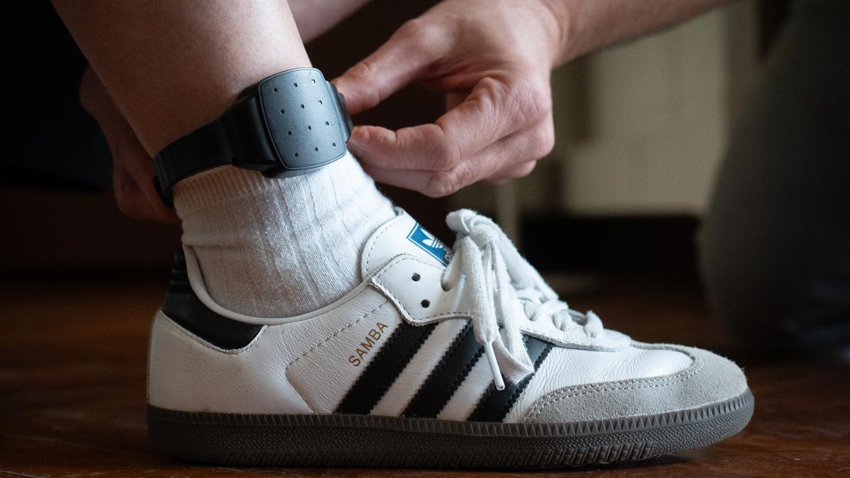 Una persona le coloca a otra una pulsera electrónica de geolocalización telemática.