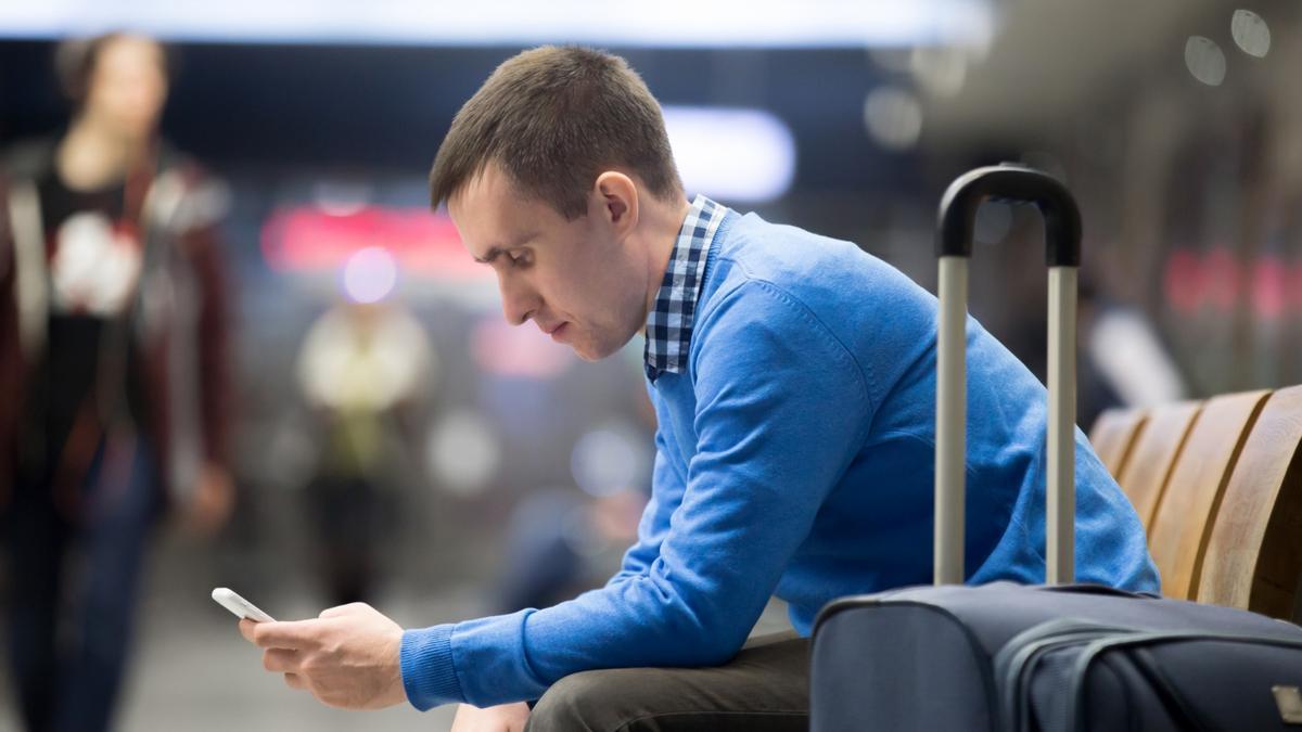 Un joven espera en un aeropuerto con su maleta y el teléfono móvil.