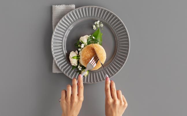 Una persona cortando una hamburguesa con cuchillo y tenedor sobre un plato. FREEPIK