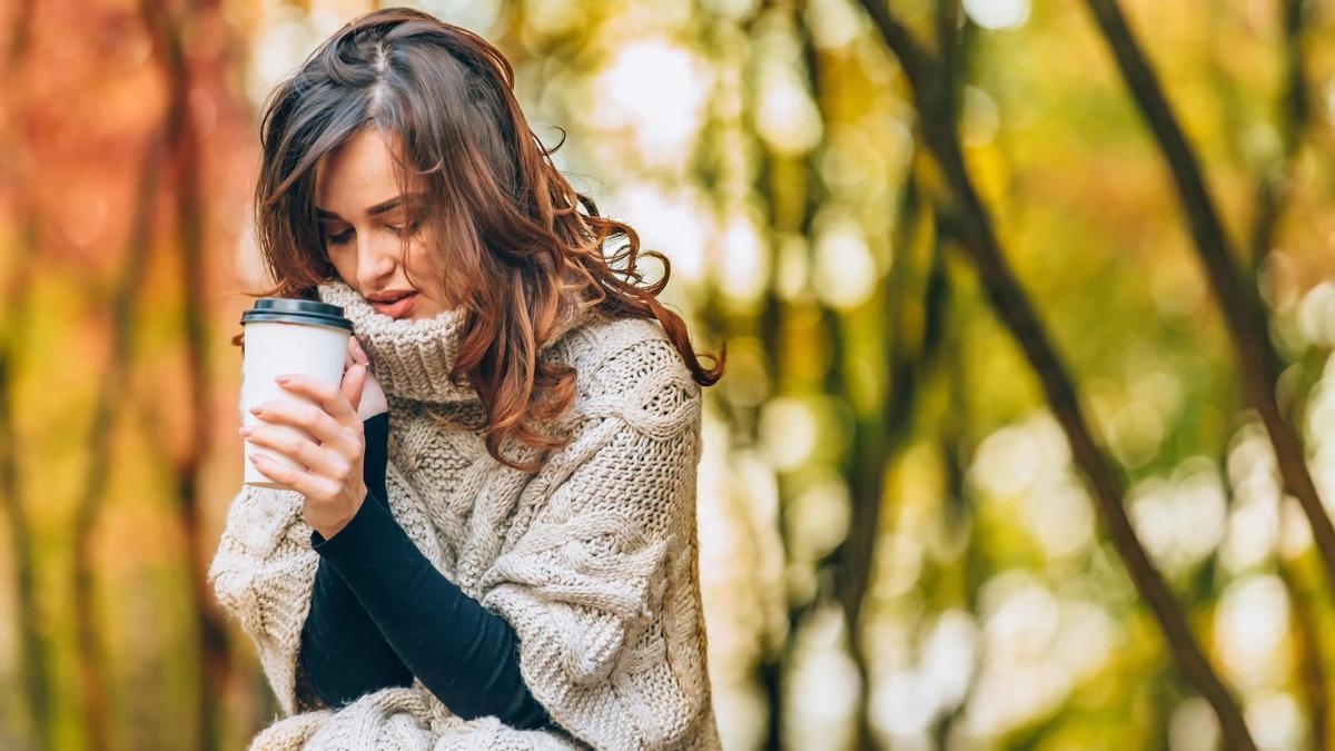 Una mujer da muestras de apatía mientras toma un café sola en un paisaje otoñal.