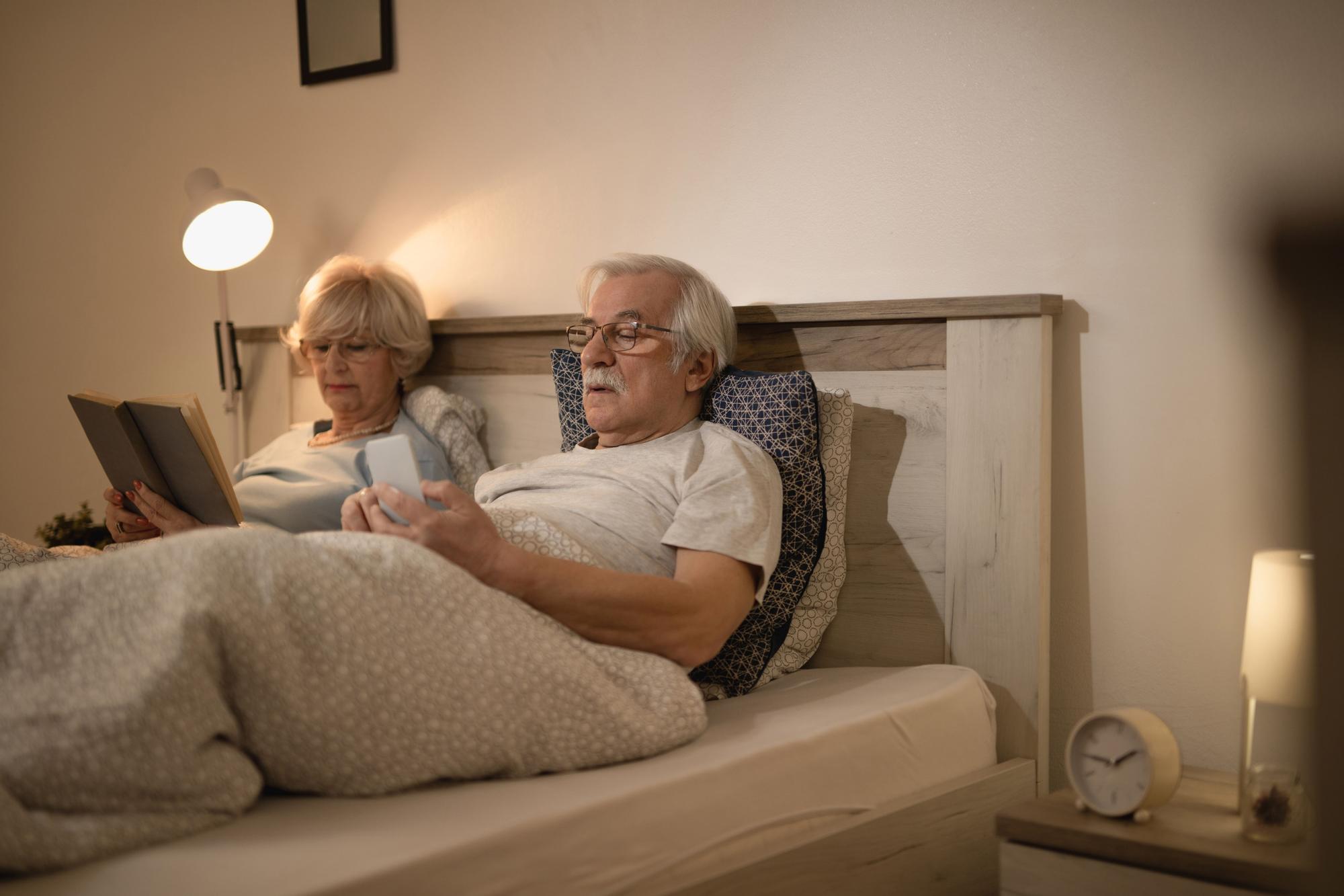 Matrimonio de personas mayores leen en la cama antes de dormir con luz fácilmente accesible en las mesillas de noches.