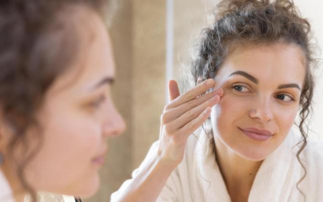 Mujer se aplica crema en la cara mientras se mira en el espejo.