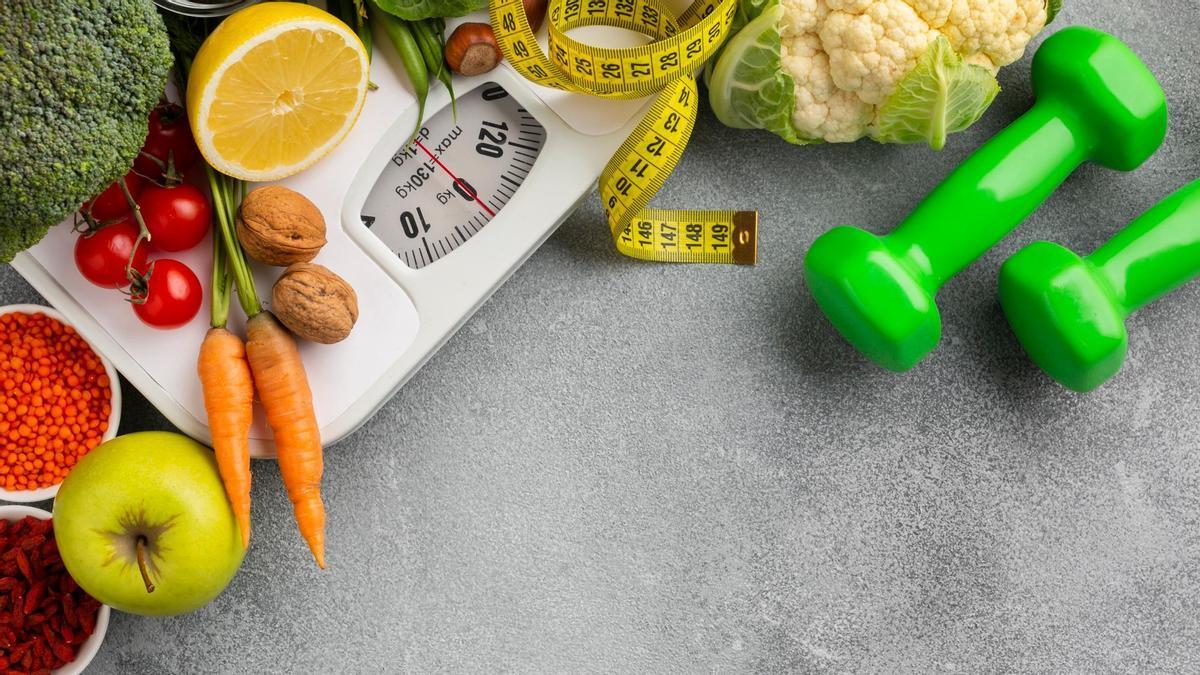Una báscula y un metro rodeados de mancuernas y alimentos saludables.