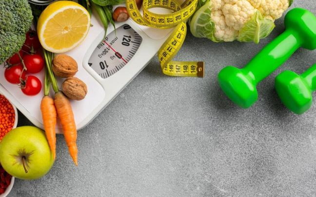 Una báscula y un metro rodeados de mancuernas y alimentos saludables. Una báscula y un metro rodeados de mancuernas y alimentos saludables.
