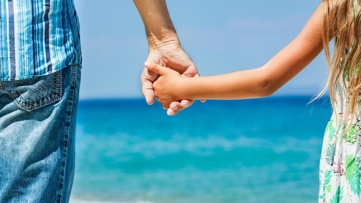 Niña de la mano de su padre en la playa con el mar de fondo.
