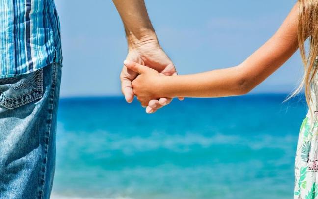 Niña de la mano de su padre en la playa con el mar de fondo.