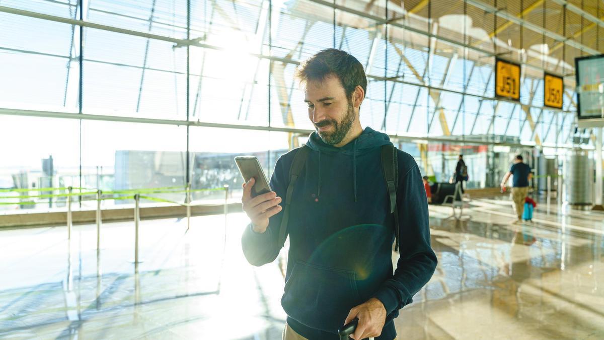 Un hombre camina por un aeropuerto con la maleta y el móvil en la mano.