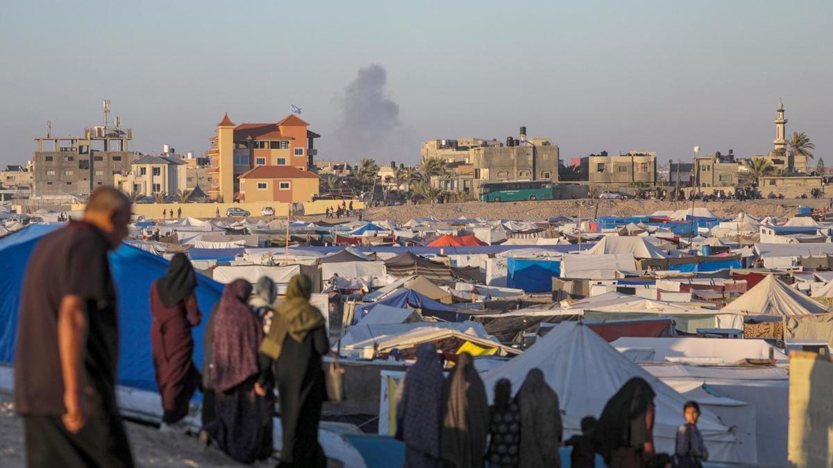 Desplazados palestinos permanecen en un campamento mientras se eleva humo al fondo tras un ataque aéreo israelí en el sur de Rafah.