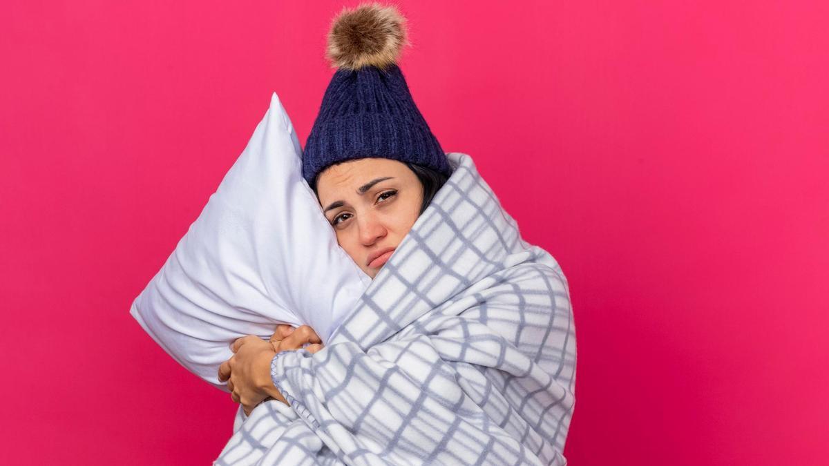 Una mujer con gorro de lana y envuelta en una manta se aferra a la almohada.