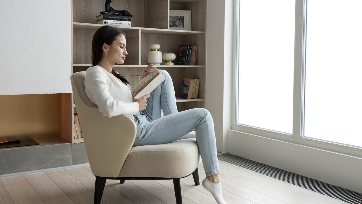 Chica lee un libro junto a la ventana sentada en el sillón.