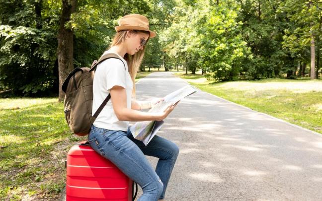 Una mujer, sentada sobre su maleta, consulta un mapa en un parque.