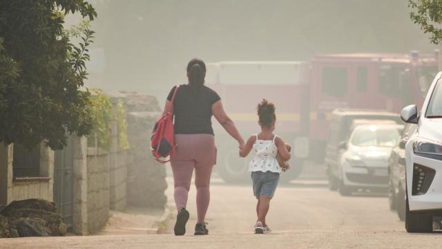 Una madre y una niña camina de la mano en Burgohondo (Ávila) entre el humo del incendio que continúa fuera de la capacidad de extinción.