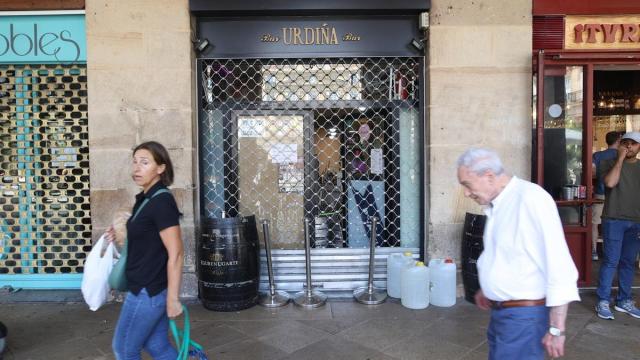 Varias personas caminan frente a una zona de bares y restaurantes con las persianas echadas.
