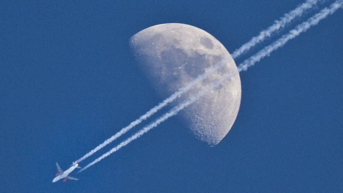Un avión deja su estela en el cielo, ante la luna en cuarto creciente.