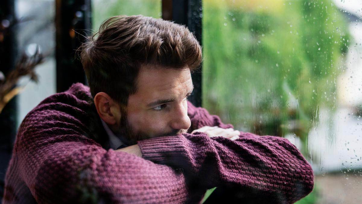 Un hombre desmotivado mira por una ventana en un día de lluvia.