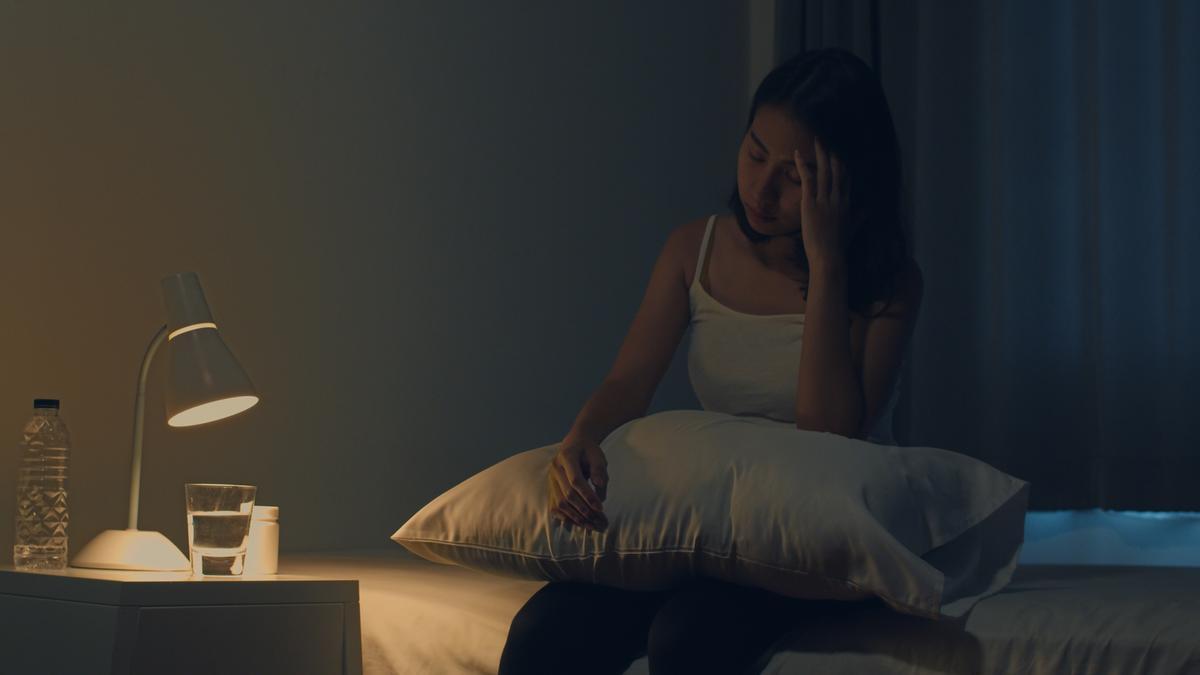 Chica sentada en la cama con la habitación a oscuras y una pequeña luz encendida en su mesilla de noche.