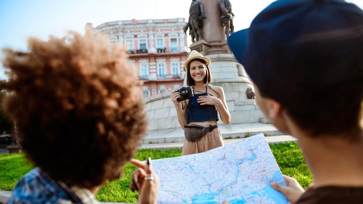 Dos personas de espaldas con u mapa y una chica sonriente que lleva una cámara de fotos para inmortalizar los mejores momentos de su viaje
