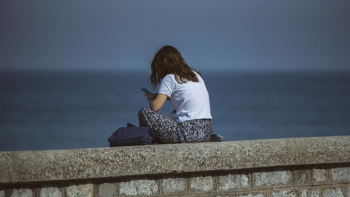 Una chica sentada sola frente al mar mirando su móvil.