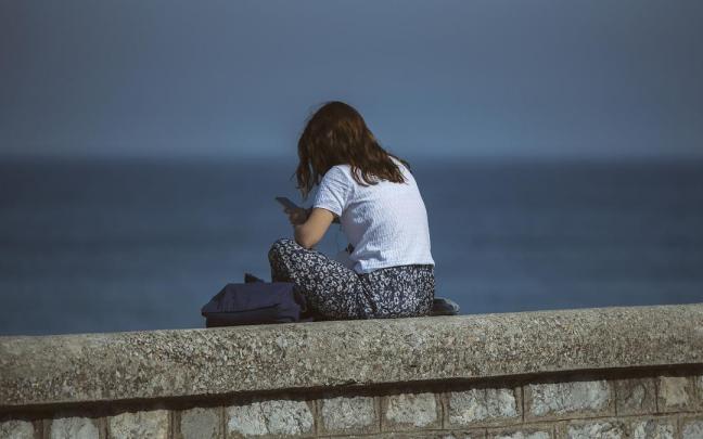 Una chica sentada sola frente al mar mirando su móvil.
