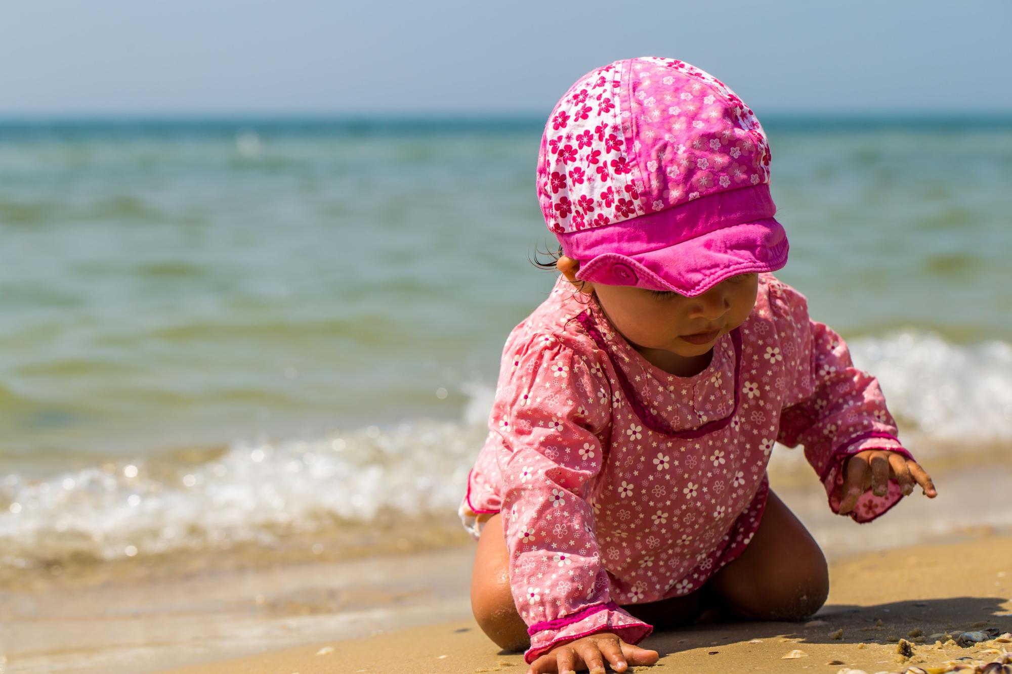Bebé en la playa con un bañador rosa de flores y una gorrita para protegerse del sol.