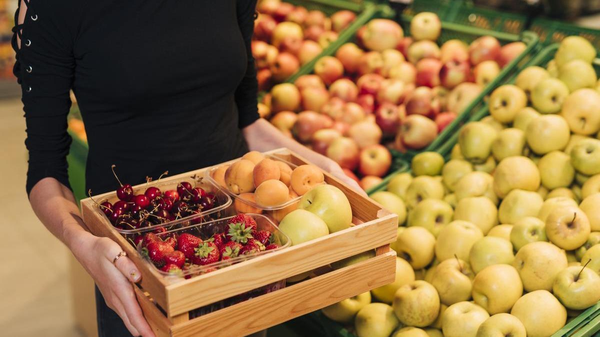 Chica en una frutería sostiene una cesta de madera con fresas, cerezas, manzanas y melocotones.