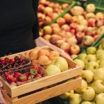 Chica en una frutería sostiene una cesta de madera con fresas, cerezas, manzanas y melocotones.
