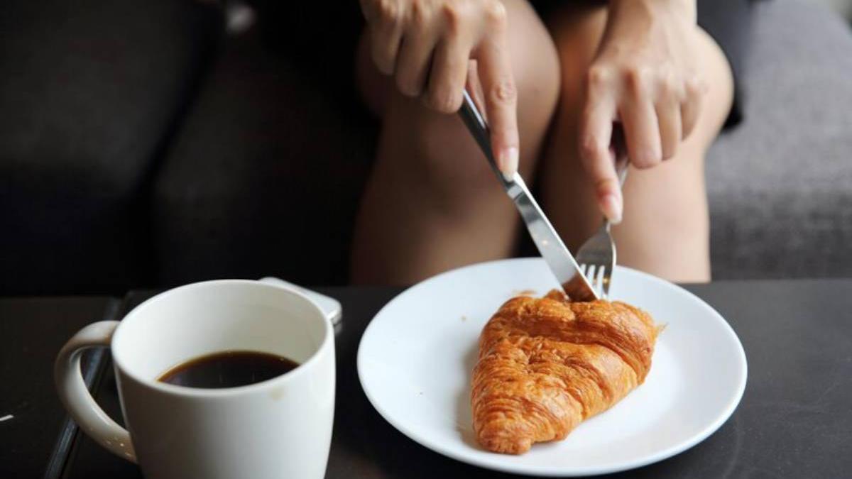 Chica desayuna un croissant acompañado de un café.