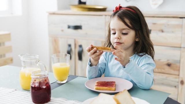 Niña desayuna una tostada con mermelada de fresa y zumo de naranja antes de ir al colegio.