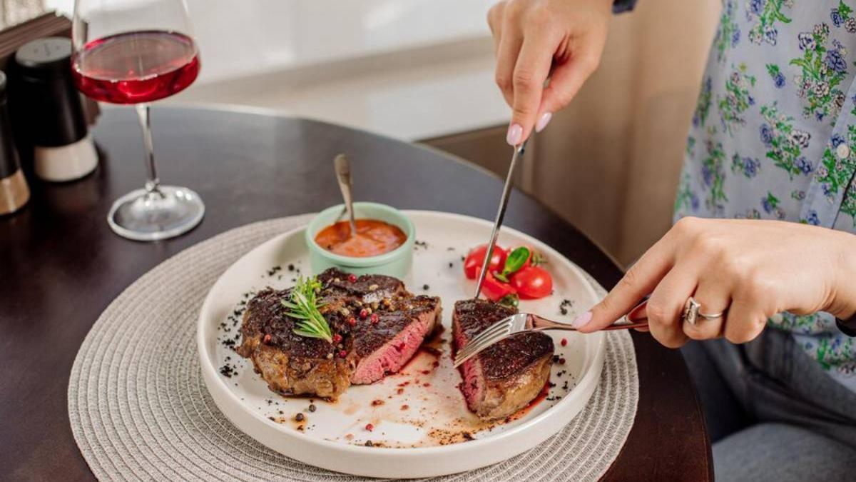 Mujer disfruta de un plato de carne y acompaña la comida con una copa de vino.