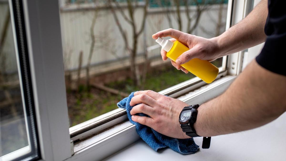 Una persona limpia con una bayeta y un spray el riel de una ventana.