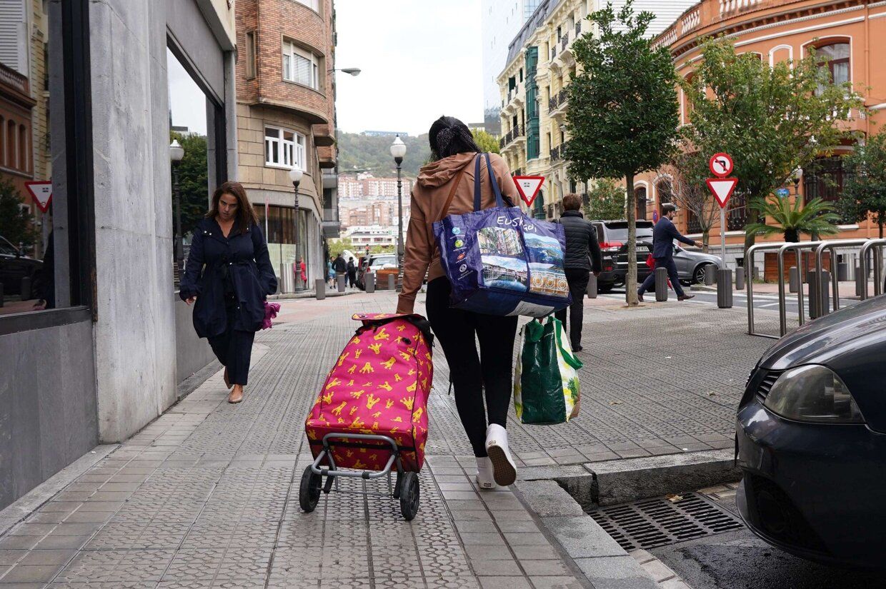 Una mujer camina cargada con varias bolsas y un carrito de la compra.