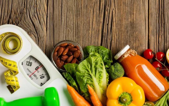 Alimentos saludables junto a una báscula, una cinta de sastre y una mancuerna.