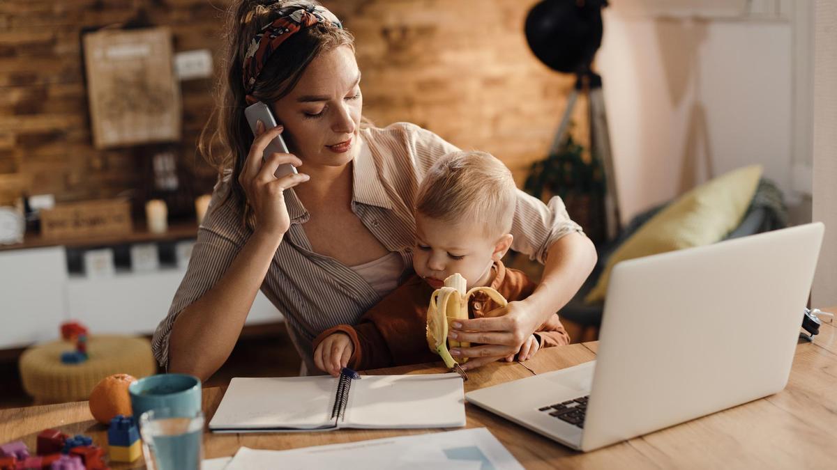 Una mujer trabaja y hace una llamada en casa mientras da la merienda a su hijo.