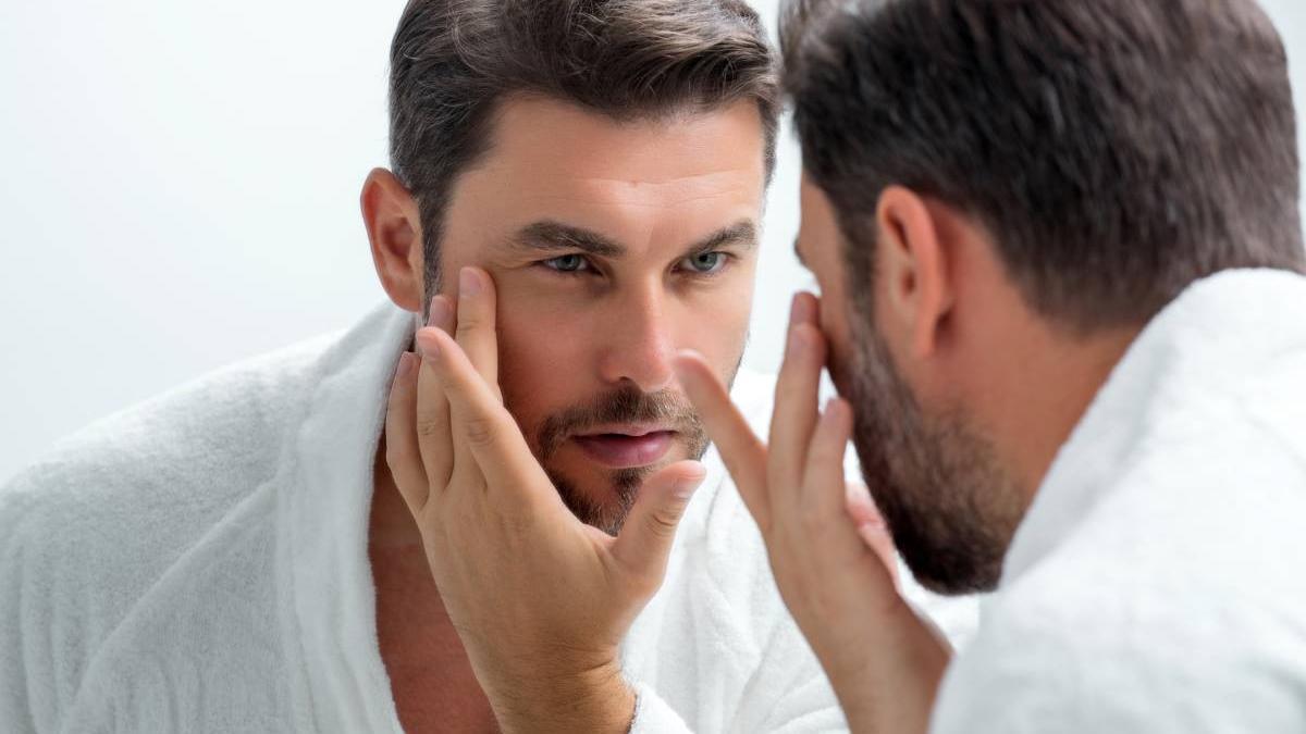 Hombre observa su rostro frente al espejo para comprobar el estado de su piel.