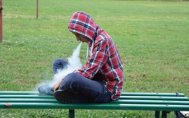 Un joven vapea en el banco de un parque.