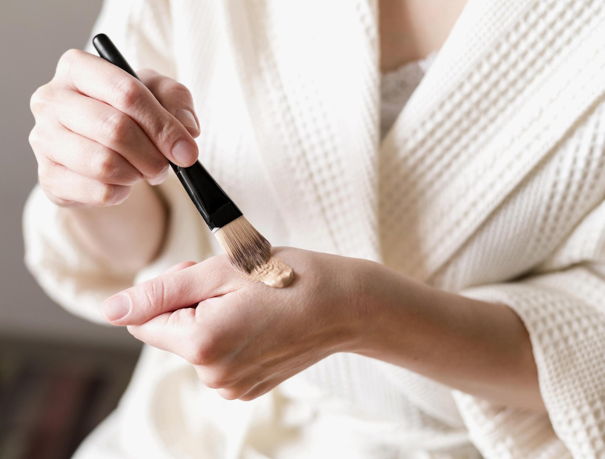 Chica aplica una pequeña cantidad de crema hidratante con color en la mano y procede a extenderla en la piel con ayuda de una brocha.