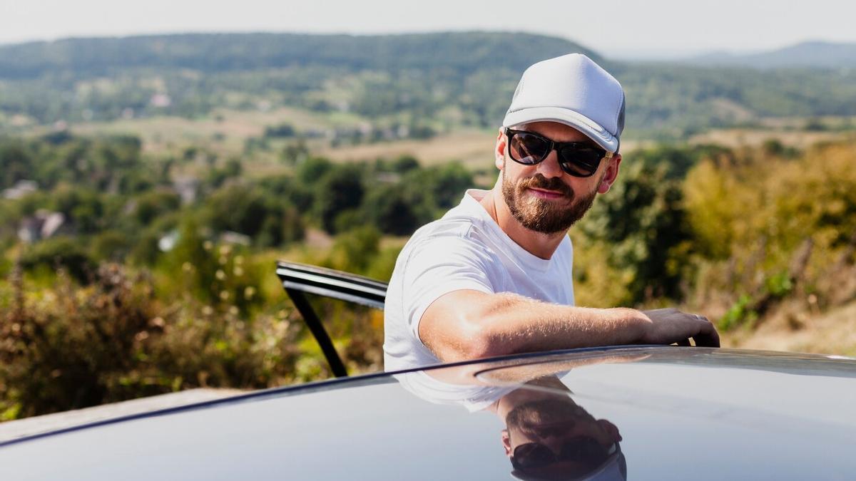 Hombre con gafas de sol y gorras apoyado en el coche.