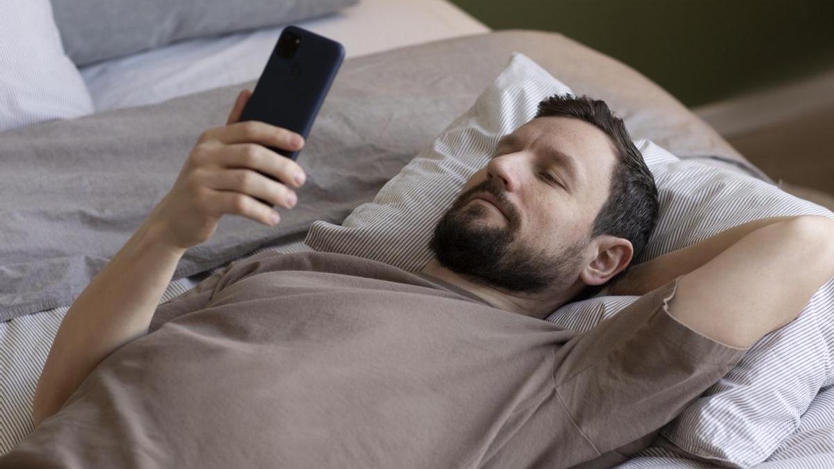 Hombre mira el móvil tumbado en la cama.