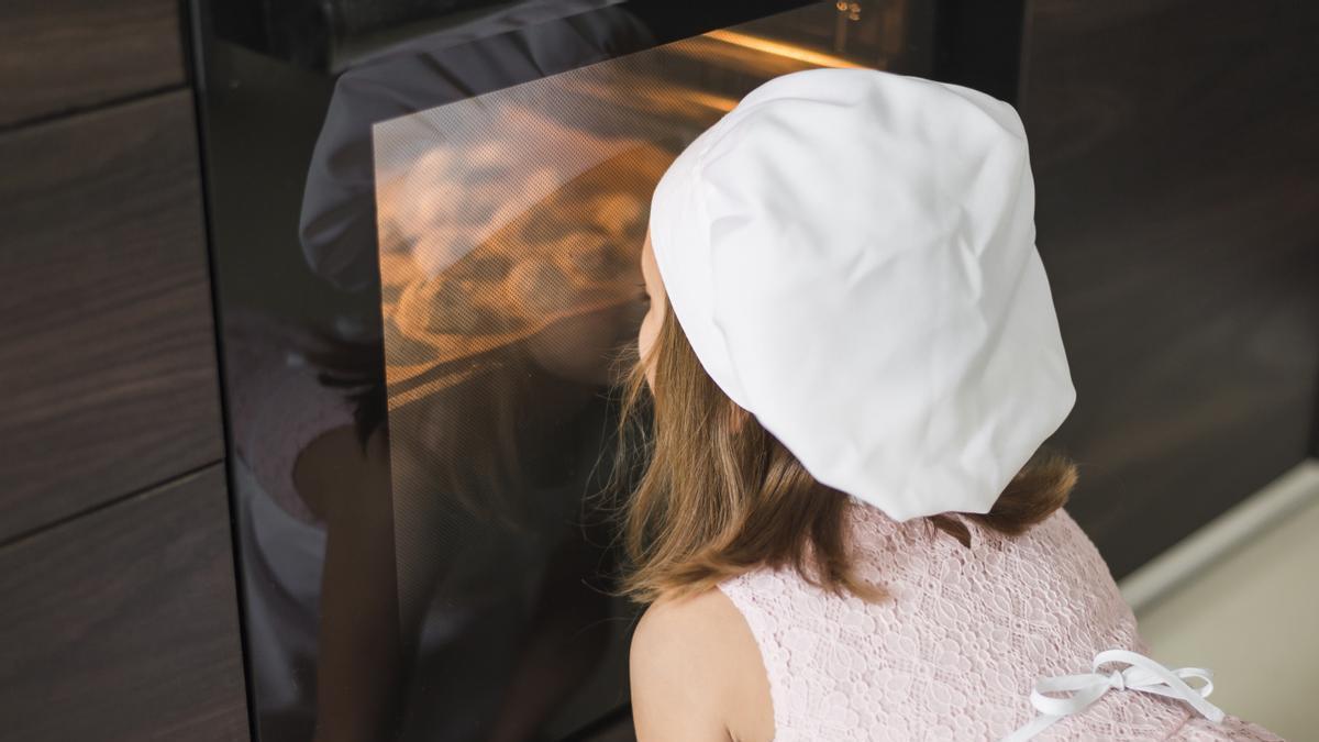 Una niña observa atentamente las galletas caseras que acaba de preparar, mientras se hornean a través de la puerta del horno.