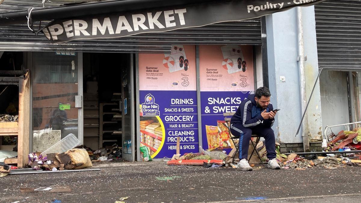 Un hombre se sienta ante su supermercado, destruido durante los disturbios en Belfast en 2024.
