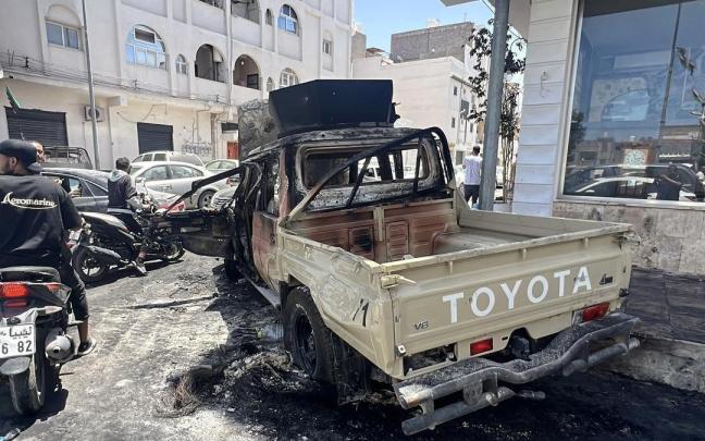 Varias personas pasean en moto ante un coche destrozado durante los ataques en Trípoli.