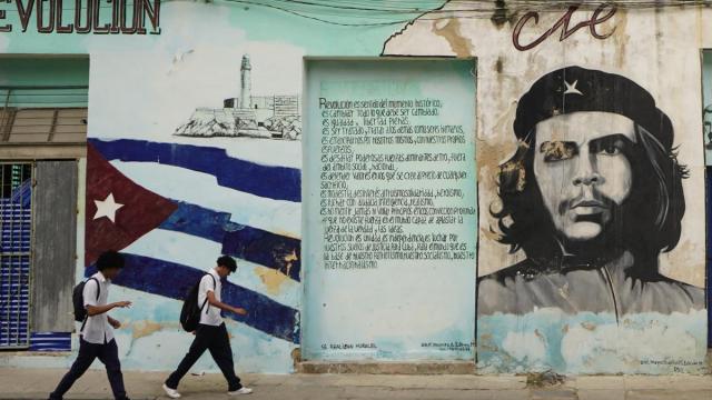Dos jóvenes pasean por las calles de La Habana junto a una bandera cubana y un mural del Che Guevara.