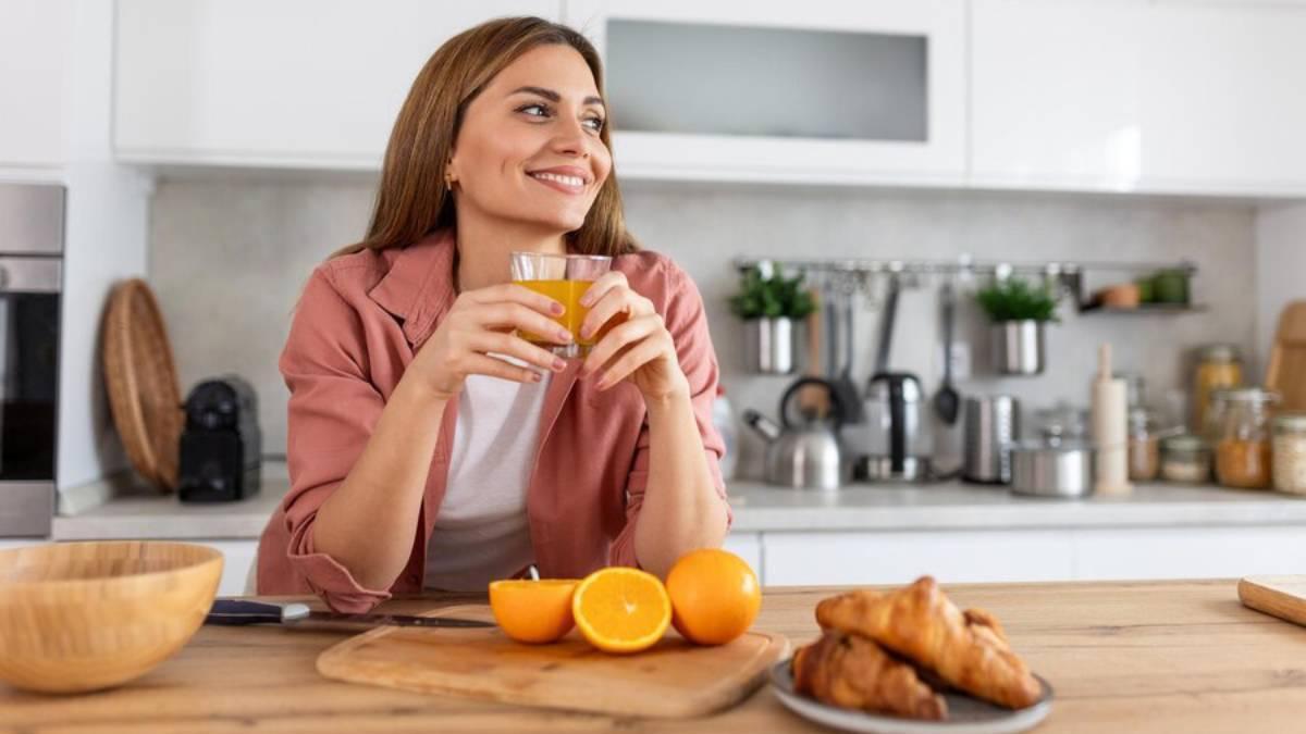 Mujer sonriente desayuna zumo de naranja recién exprimido en la encimera de su cocina