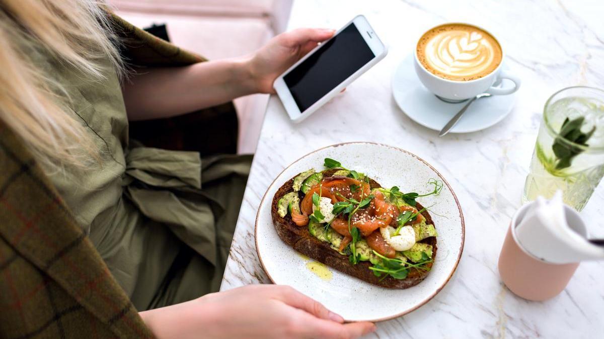 Chica sentada en una terraza desayuna una tostada de aguacate y tomate con un café con leche mientras mira el móvil