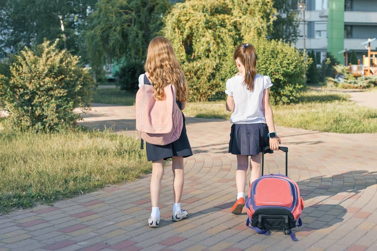 Dos niñas van al colegio, una de ellas lleva una mochila a la espalda y otra una con ruedas.