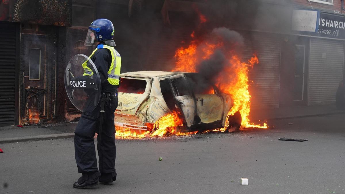 Un policía, junto a un coche en llamas a causa de los disturbios registrados el pasado fin de semana en Reino Unido por el apuñalamiento mortal de tres niñas.