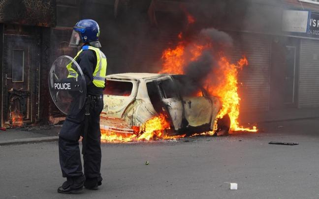 Un policía, junto a un coche en llamas a causa de los disturbios registrados el pasado fin de semana en Reino Unido por el apuñalamiento mortal de tres niñas.