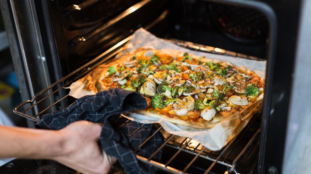 Manos de un hombre sacando la pizza a base de verduras del horno con ayuda de un trapo de cocina para no quemarse