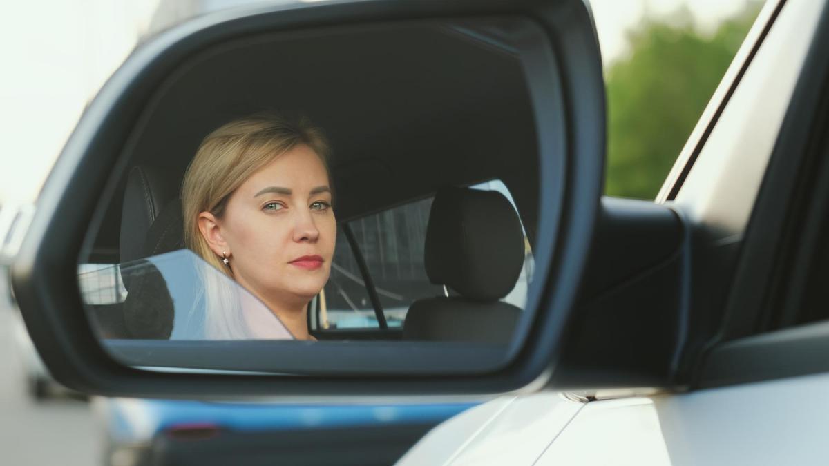 Una mujer reflejada en el espejo retrovisor lateral izquierdo.